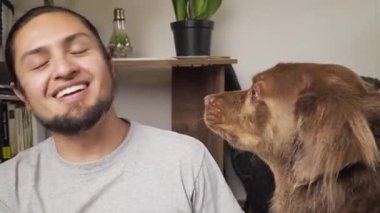 Kahverengi köpek bir adamın burnunu yalayarak şefkat gösteriyor, ikisi de kapalı bir ortamda hassas bir anı paylaşıyor..