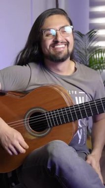 Uzun saçlı ve gözlüklü neşeli bir adam evde rahat bir ortamda otururken klasik gitar çalar..