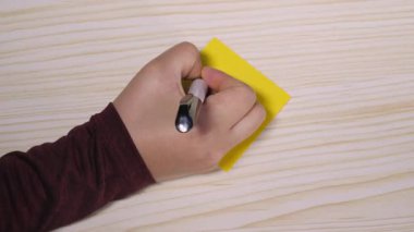 Tahta bir masa yüzeyindeki sarı yapışkan bir nota yazı yazarken elinde siyah bir kalem vardı..