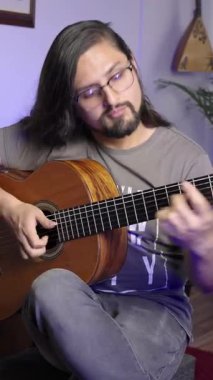 Uzun saçlı ve gözlüklü bir müzisyen klasik gitar çalar, konsantrasyon ve müzik tutkusu gösterir..