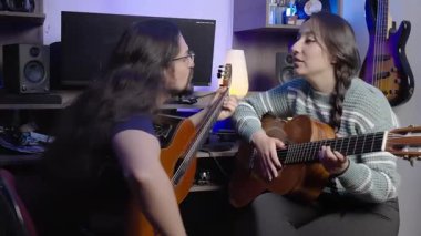 Arka planda enstrümanlar ve ses ekipmanları olan samimi bir müzik stüdyosunda akustik gitarlarla öpüşen iki kişi..