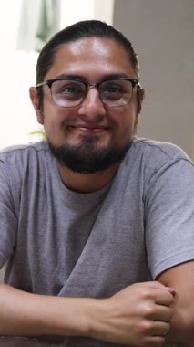 Gülümseyen bir adamın portresi, gözlüklü, sakallı, sırtı bağlı, gri tişörtlü, kapalı bir ortamda..