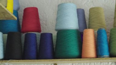 Kırmızı, mavi, yeşil, turuncu ve sarı gibi çeşitli renklerde iplik külahları ahşap bir rafta beyaz desenli bir duvara karşı düzgünce dizilmiş..