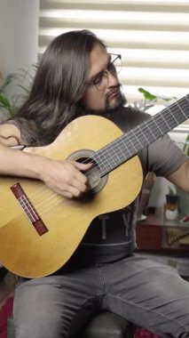 Uzun saçlı ve gözlüklü bir adam klasik gitar çalıyor. Arka planda bitkiler olan kapalı bir ortamda oturuyor..
