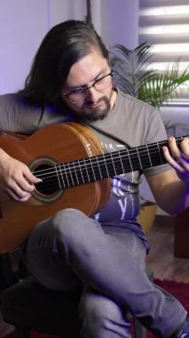 Gözlüklü ve uzun saçlı genç adam içeride ahşap akustik gitar çalıyor, müziğine odaklanmış..
