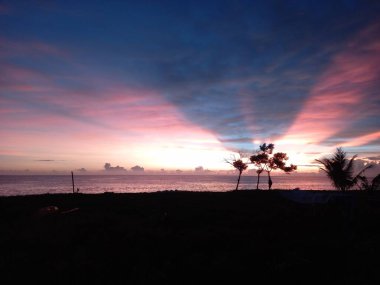 Gün doğumunda plajda, kırmızı gökyüzü ve ağaç siluetleri.