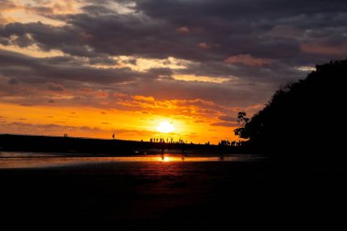 Sahildeki siluetleri dramatik turuncu bir gün batımına karşı bulut oluşumları ve okyanus yansımalarıyla.
