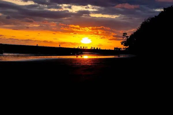 Sahildeki siluetleri dramatik turuncu bir gün batımına karşı bulut oluşumları ve okyanus yansımalarıyla.