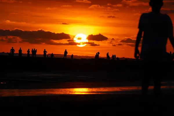 Sahildeki siluetleri dramatik turuncu bir gün batımına karşı bulut oluşumları ve okyanus yansımalarıyla.