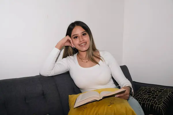 Evdeki kanepede oturmuş kitap okuyan genç bir kadın.