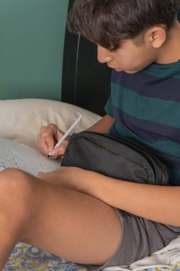 Genç bir adam siyah bir çanta ve kalemle bir yatakta oturuyor.