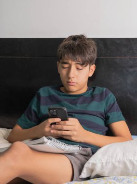 Genç çocuk yatakta defterle ders çalışırken telefon kullanıyor.