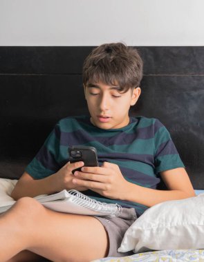 Genç çocuk yatakta çalışırken telefonla dikkatini dağıtıyor.