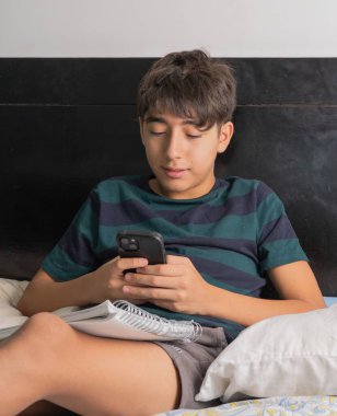 Genç bir adam yatakta oturuyor ve cep telefonuna bakıyor.