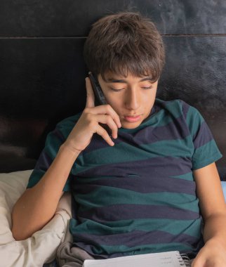 Genç bir adam yatakta otururken cep telefonuyla konuşuyor.