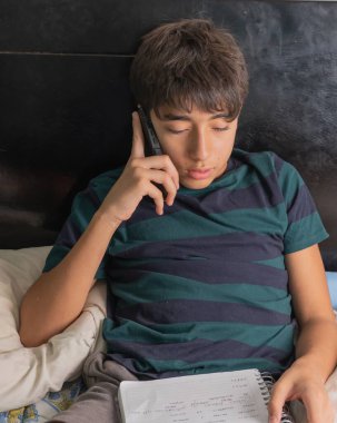 Genç çocuk yatakta çalışırken telefonla dikkatini dağıtıyor.