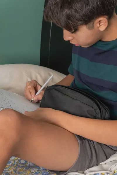 Genç bir adam siyah bir çanta ve kalemle bir yatakta oturuyor.