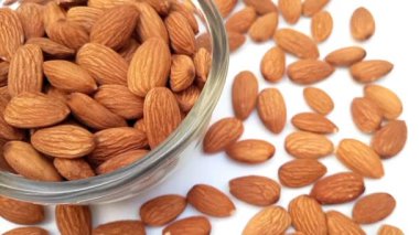 Bademler cam bir kasede, beyaz arka plan köşesinde dönüyor ve bademler her yere yayılıyor. Kabuksuz badem, kapat