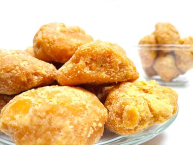 Arka planda bir kase dolusu organik kahverengi kamış şekeri olan temiz beyaz bir zemin üzerinde cam bir tabakta Jaggery veya Gur parçaları, yakın plan.