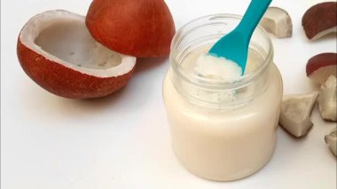 Kavanozdaki sert hindistan cevizi yağının yanında beyaz arka planda beyaz hindistan cevizi kırıkları olan plastik bir kaşık. Kapat.