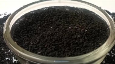 Siyah kimyon tohumları veya siyah kimyon tohumları beyaz zemin üzerinde dönen bir cam kaseye düşer. Etrafına baharatlar saçılmıştır. Mikro çekim, merkez çerçeve, otlar ve baharatlar.