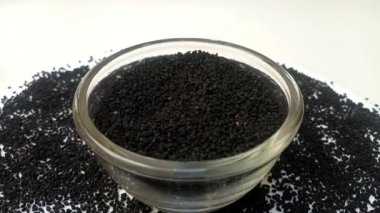 Siyah kimyon tohumları veya cam bir kaptaki siyah kimyon tohumları etrafa saçılmış, beyaz arka plan, merkez çerçeve, yakınlaşma, kapanış, bitkiler ve baharatlar üzerinde dönüyor