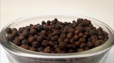 Beyaz arka planda bir cam kasede siyah biber damlaları, ortada dönen siyah biberlerle dolu bir cam kase, mikro shot, bitkiler ve baharatlar..