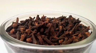 Karanfiller beyaz arka planda cam bir kaseye düşer, merkezde karanfil dolu bir cam kase döner, mikro çekim, otlar ve baharatlar.