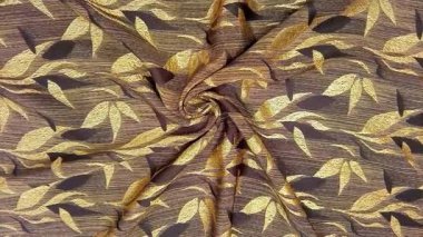 Tekstil kumaş hareketli arka planda kahverengi ve altının çeşitli tonlarında tekrarlanan yapraklar var. Çiçek veya yaprak benzeri motifler simetrik desenlerde sıralanmış, basitleştirilmiş.