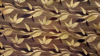 Tekstil kumaş hareketli arka planda kahverengi ve altının çeşitli tonlarında tekrarlanan yapraklar var. Çiçek veya yaprak benzeri motifler simetrik desenlerde sıralanmış, basitleştirilmiş.