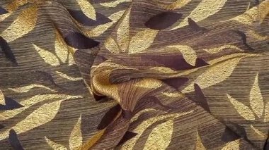 Tekstil kumaş hareketli arka planda kahverengi ve altının çeşitli tonlarında tekrarlanan yapraklar var. Çiçek veya yaprak benzeri motifler simetrik desenlerde sıralanmış, basitleştirilmiş.