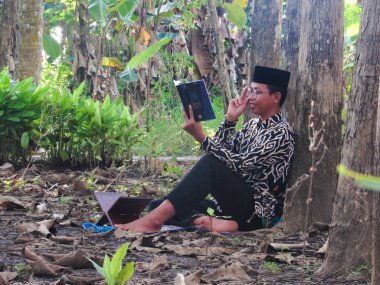 Asyalı Müslüman bir adam oturuyor ve Kuran okuyor. Ormanın ortasındaki barış onu dindar ve geleneksel bir atmosfer haline getiriyor.