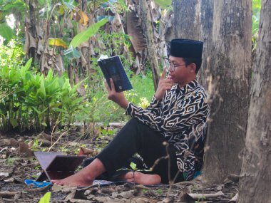 Asyalı Müslüman bir adam oturuyor ve Kuran okuyor. Ormanın ortasındaki barış onu dindar ve geleneksel bir atmosfer haline getiriyor.