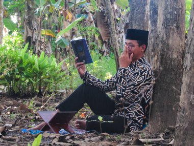 Asyalı Müslüman bir adam oturuyor ve Kuran okuyor. Ormanın ortasındaki barış onu dindar ve geleneksel bir atmosfer haline getiriyor.