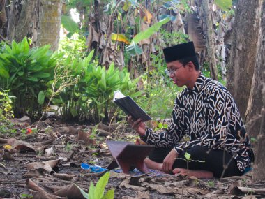 Asyalı Müslüman bir adam oturuyor ve Kuran okuyor. Ormanın ortasındaki barış onu dindar ve geleneksel bir atmosfer haline getiriyor.