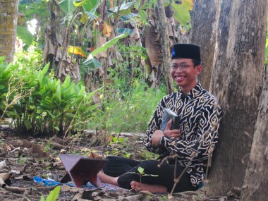 Asyalı Müslüman bir adam oturuyor ve Kuran okuyor. Ormanın ortasındaki barış onu dindar ve geleneksel bir atmosfer haline getiriyor.