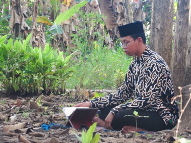 Asyalı Müslüman bir adam oturuyor ve Kuran okuyor. Ormanın ortasındaki barış onu dindar ve geleneksel bir atmosfer haline getiriyor.