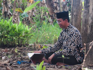 Asyalı Müslüman bir adam oturuyor ve Kuran okuyor. Ormanın ortasındaki barış onu dindar ve geleneksel bir atmosfer haline getiriyor.