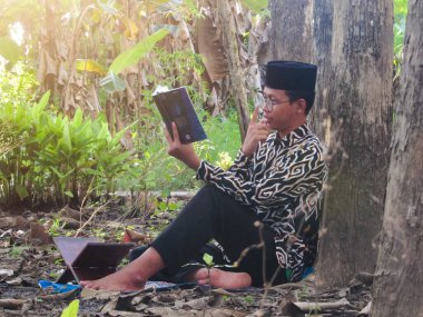 Asyalı Müslüman bir adam oturuyor ve Kuran okuyor. Ormanın ortasındaki barış onu dindar ve geleneksel bir atmosfer haline getiriyor.