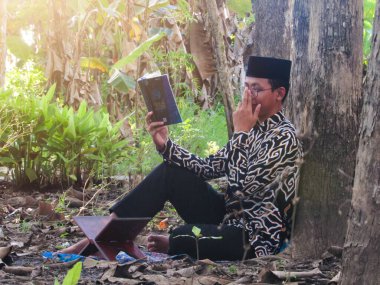 Asyalı Müslüman bir adam oturuyor ve Kuran okuyor. Ormanın ortasındaki barış onu dindar ve geleneksel bir atmosfer haline getiriyor.