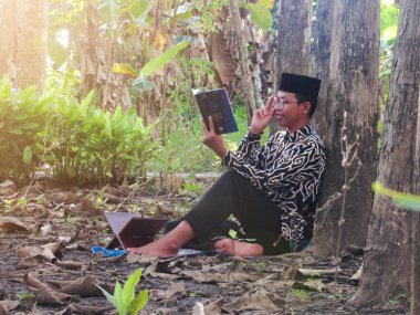 Asyalı Müslüman bir adam oturuyor ve Kuran okuyor. Ormanın ortasındaki barış onu dindar ve geleneksel bir atmosfer haline getiriyor.