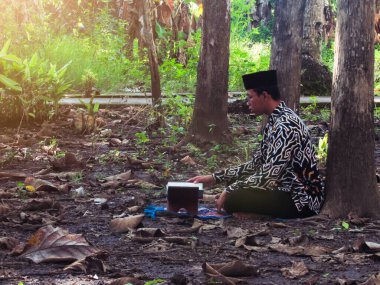 Asyalı Müslüman bir adam oturuyor ve Kuran okuyor. Ormanın ortasındaki barış onu dindar ve geleneksel bir atmosfer haline getiriyor.