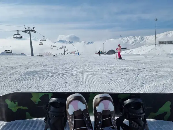 Ön planda kamuflaj desenli snowboard. Karla kaplı dağlar, kayakçılar ve bulutlu bir gökyüzünün altında kayak asansörleri. Açık hava aktiviteleri için mükemmel bir kış manzarası.