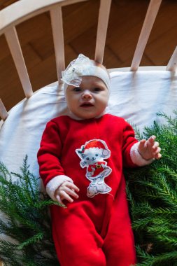 A small baby in a bright red Christmas outfit lying in a round wooden cradle. Festive atmosphere at home. clipart