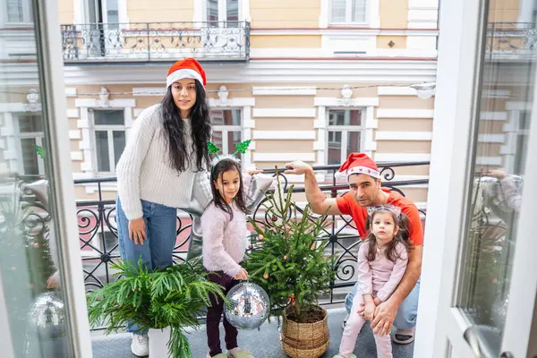Baba ve kızları balkonda küçük bir Noel ağacı ve şenlik alanı süslüyorlar. Yeni yıl atmosferi, parlak süslemeler ve mutlu gülümsemeler ailede neşeli bir tatil öncesi havası yaratır..