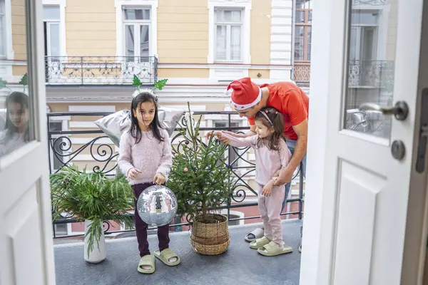 Baba ve kızları balkonda küçük bir Noel ağacı ve şenlik alanı süslüyorlar. Yeni yıl atmosferi, parlak süslemeler ve mutlu gülümsemeler ailede neşeli bir tatil öncesi havası yaratır..