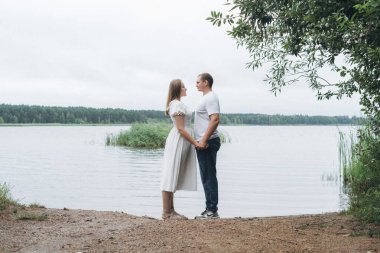 Loving couple shares intimate moments in nature near a scenic lake and forest. They hug, kiss, and hold hands, expressing deep emotions. The woman wears a flowing white dress, evoking a wedding elopement vibe. The couple experiences a mix of love, te clipart