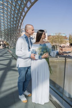 Geleneksel Ukrayna nakışlı düğün kıyafeti giymiş yeni evliler, Gürcistan 'ın Tiflis kentindeki modern cam mimarinin önünde zarif bir şekilde poz veriyorlar. Beyaz gelinlikli gelin bir buket narin mavi çiçek tutuyor. Kültürünün mükemmel bir karışımı.