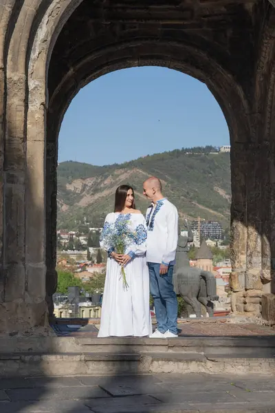 Geleneksel Ukrayna işlemeli düğün kıyafeti giyen yeni evli bir çift, Gürcistan 'ın Tiflis kentindeki antik bir kemerin altında romantik bir öpücük paylaşıyor. Gelin beyaz bir gelinlik giyiyor ve damat onu sevgiyle kucaklıyor. Bir yaz tatili düğünü.