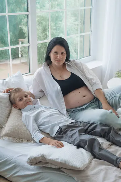 Samimi bir aile fotoğrafı, hassas bir beklenti anını yakalıyor. Siyah bluz ve düğmesiz beyaz gömlek giyen hamile anne elini nazikçe karnına koyar. Beyaz gömlekli baba, onun arkasında korunuyor.